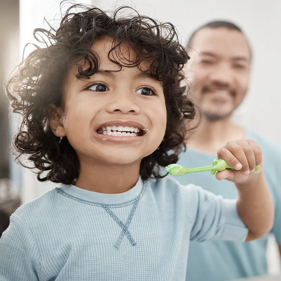 A little boy smiling brushing his teeth as his father looks on