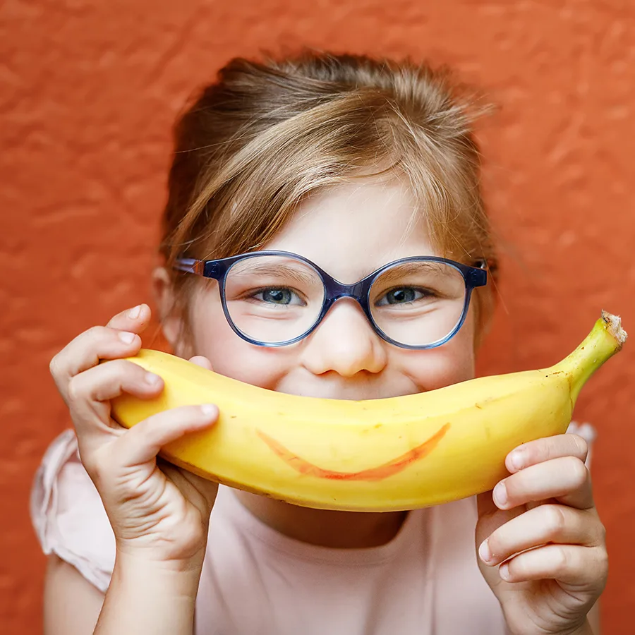 A young girl in glasses holds a banana in place of her smile